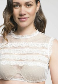 Durchsichtiges weißes Crop-Top mit Spitzenbesatz und Polka-Dot-Muster. Es hat kurze Ärmel und einen gewellten Ausschnitt, der über einem hautfarbenen Unterwäschestück liegt.