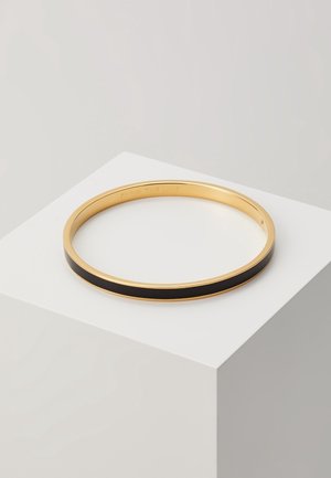 Goldener Armreif mit schwarzer Emaille-Einlage, der auf einem weißen Sockel vor einem hellgrauen Hintergrund liegt.