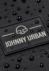 Fekete textúrájú anyag vízcseppekkel, amelyen egy márkás gumiragasztó található, melyen világos színű felirattal olvasható a "JOHNNY URBAN".