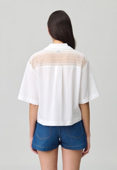 Camicetta bianca a maniche corte con pannello all'uncinetto sul retro. La camicia presenta un colletto e una vestibilità comoda, abbinata a pantaloni corti di denim.