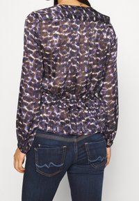 Blusa trasparente viola e nera con vita arricciata, maniche lunghe e motivo astratto; indossata con jeans in denim scuro con tasche decorative.