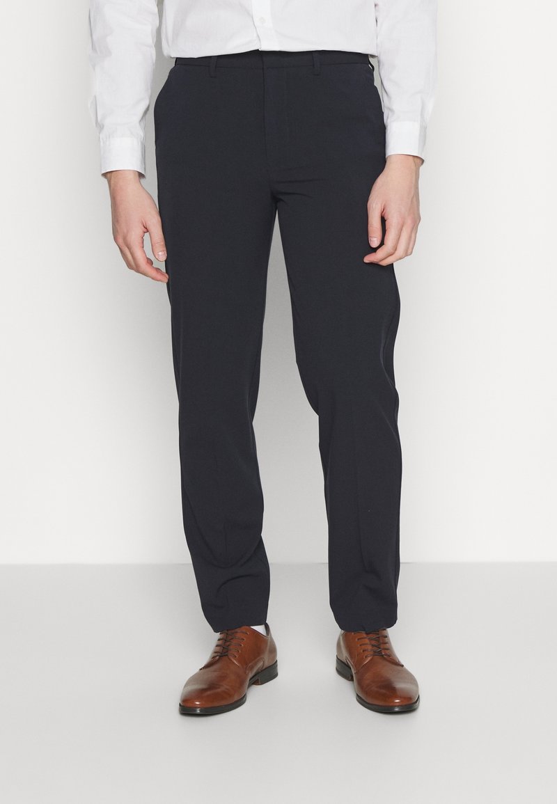 Homme portant un pantalon formel noir, une chemise blanche et des chaussures habillées en cuir marron, debout sur un sol gris.