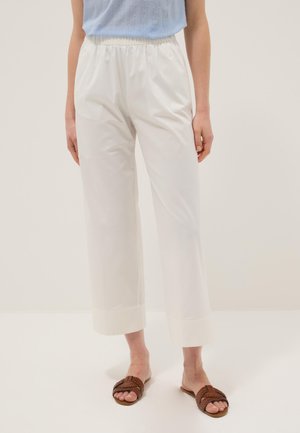 Donna che indossa pantaloni bianchi a gamba larga con orli risvoltati e sandali piatti intrecciati marroni, in piedi davanti a uno sfondo chiaro e uniforme.