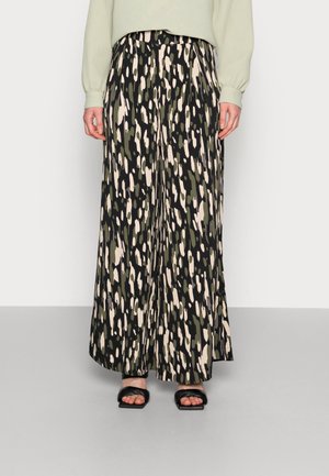 Persona con pantalones de pierna ancha con estampado abstracto en negro, verde y beige combinados con sandalias negras de tacón con punta abierta y top de punto verde claro.