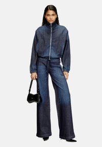 Veste en denim avec col montant et fermeture éclair, assortie à un pantalon en denim à jambes larges, le tout en bleu foncé. Tient un petit sac à main noir.