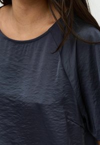 Top blu navy caratterizzato da una texture liscia e leggermente increspata, scollatura tonda ampia e dettaglio con tasca laterale. Maniche corte.