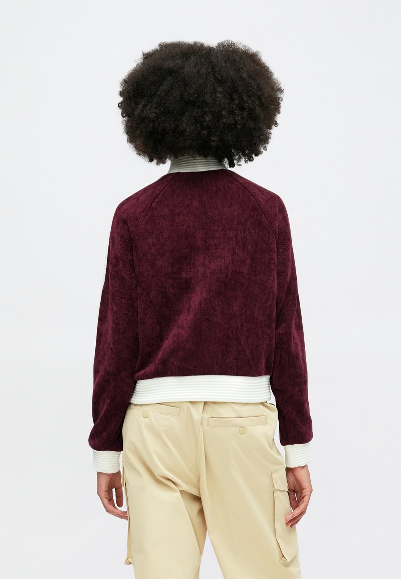 Bordeaux katoenen ribfluweel jasje met geribde witte manchetten en kraag. Heeft een relaxte pasvorm en raglanmouwen. Draagt een lichte beige broek.