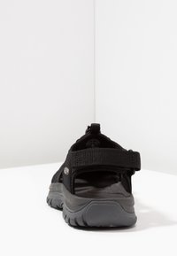 Black sandal featuring a textile upper, adjustable Velcro strap, and a thick gray rubber sole with textured tread for grip.