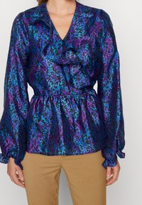 Modelka nosi jaskrawą bluzkę z fioletowym i niebieskim wzorem w zwierzęta oraz falbankami, zestawioną z beżowymi spodniami, na jednolitym tle.