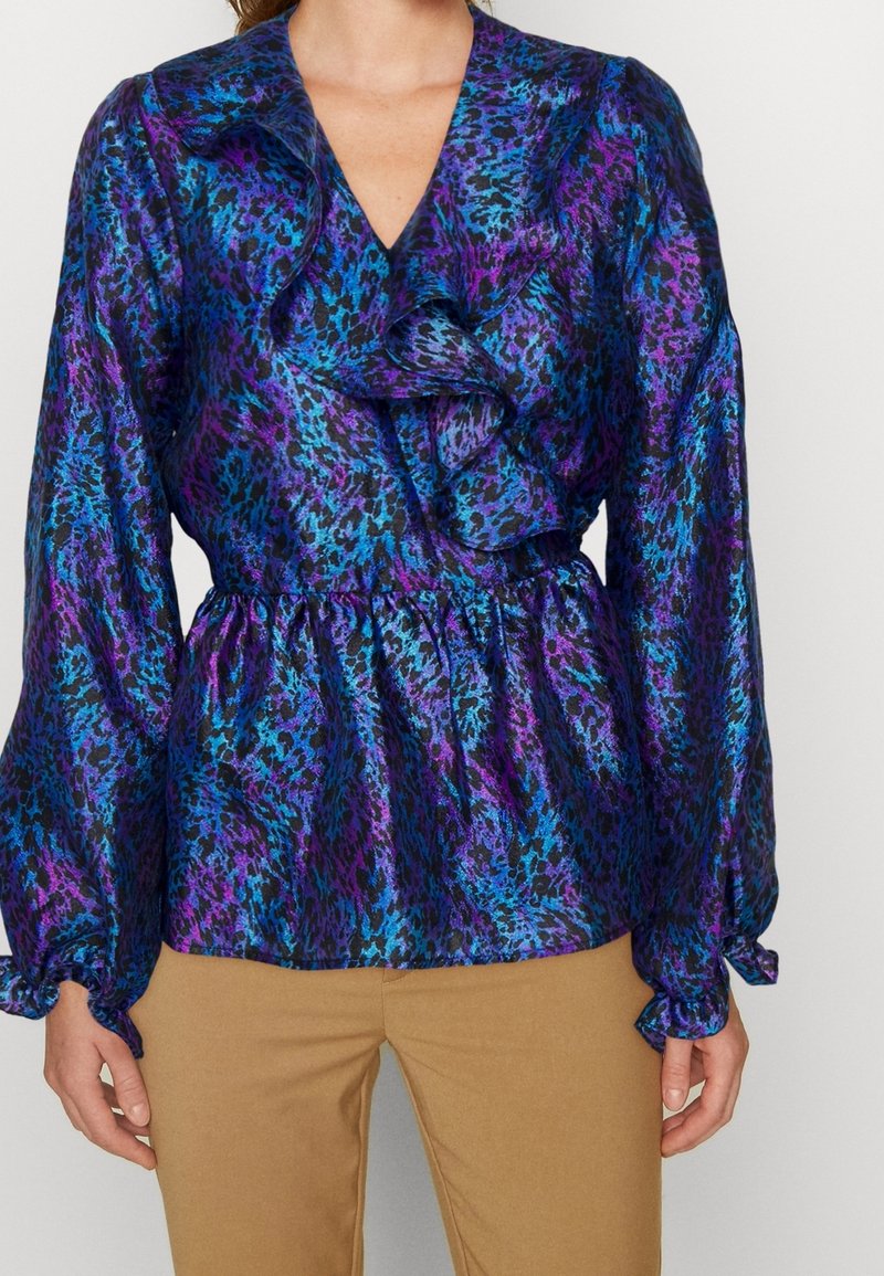 Modelka nosi jaskrawą bluzkę z fioletowym i niebieskim wzorem w zwierzęta oraz falbankami, zestawioną z beżowymi spodniami, na jednolitym tle.