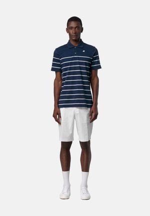 Uomo in piedi che indossa una polo a righe blu navy, pantaloncini bianchi fino al ginocchio, calzini bianchi e scarpe da ginnastica bianche su uno sfondo semplice.