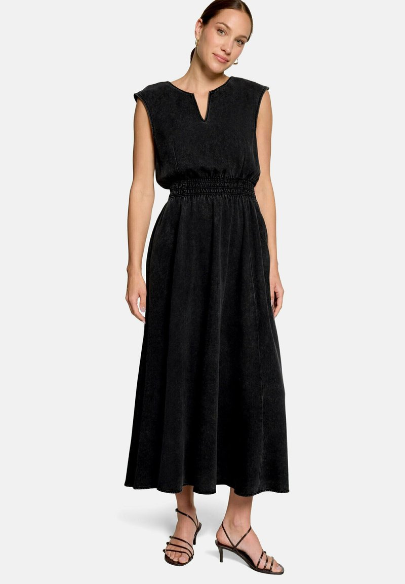 Femme portant une robe midi noire sans manches avec une taille froncée et un décolleté fendu, associée à des sandales à talons noires à lanières.