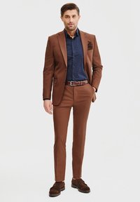 Brąswny garnitur o dopasowanym kroju, z teksturowanej tkaniny i klapami w kształcie notcha. Noszony na niebieskiej koszuli w białe kropki i brązowych butach.