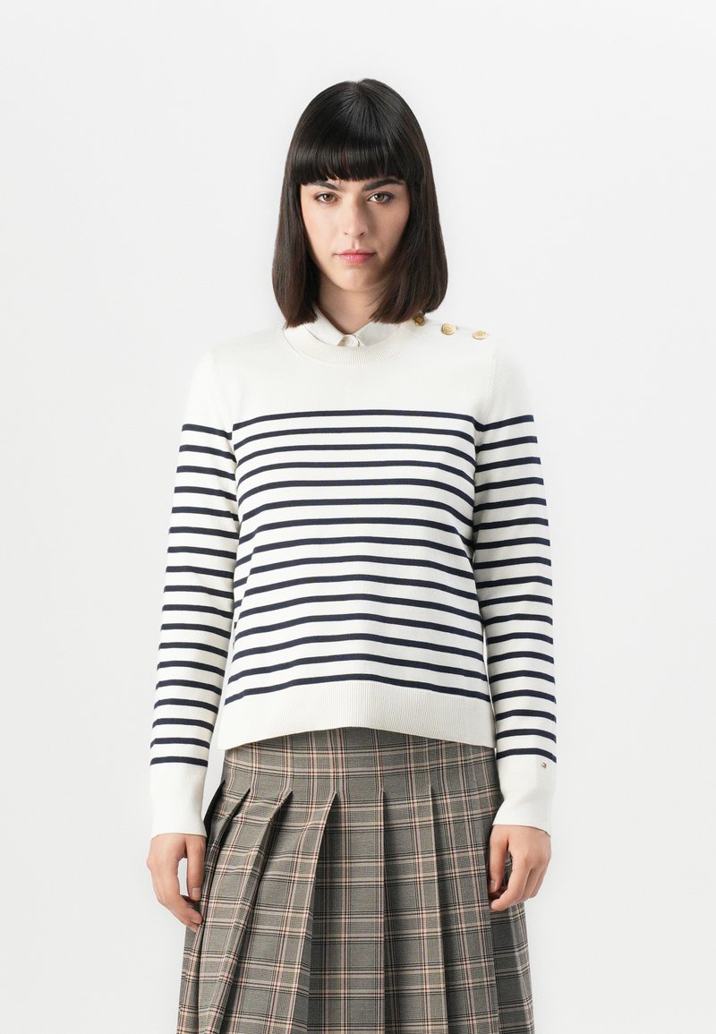 Gestreifter Strickpullover in Creme und Marineblau mit einem Kragenhals, Knopfakzenten an der Schulter und gerippten Bündchen; kombiniert mit einem karierten Rock.