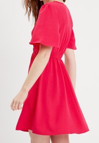 Robe rouge avec manches courtes bouffantes, taille élastique et jupe évasée. Texture lisse avec un tissu léger et un léger éclat.
