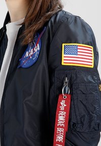 Czarna nylonowa kurtka bomberka z naszywką NASA i naszywką amerykańskiej flagi. Zawiera czerwoną metkę z napisem "USUNĄĆ PRZED", z kieszenią na zamek.
