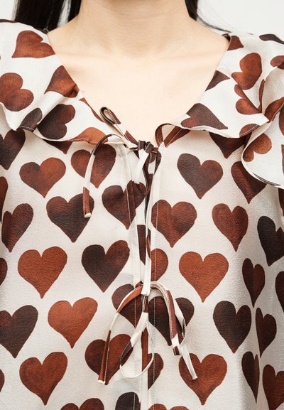 Μπλούζα με καφέ σχέδιο καρδιάς, βολάν στον γιακά και δεμένες λεπτομέρειες φιόγκων στο ντεκολτέ και το στήθος σε ανοιχτό ύφασμα.
