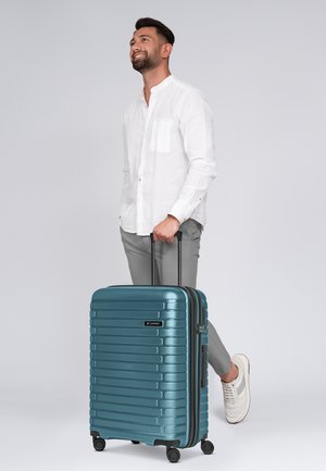 Valise rigide teal avec des rainures verticales, poignée rétractable et quatre roues pivotantes. Matériau durable avec une fermeture éclair et un détail de logo.