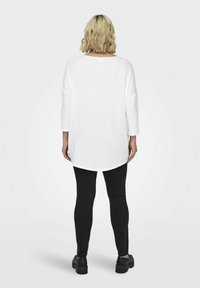 Túnica blanca de manga larga con un dobladillo redondeado, combinada con leggings negros ajustados y zapatos negros robustos. Tejido suave; corte relajado.