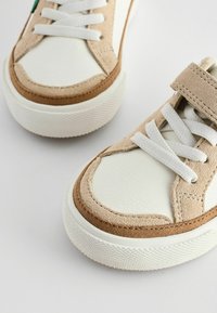 Hvide lædersneakers med tan ruskindsdetaljer, udstyret med hvide snørebånd og en velcrosnor. Struktureret gummisål.