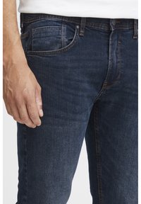 Mörkblå denimjeans med en slät textur, fem fickor och subtil urvigning. Har synlig sömnad och knappstängning.