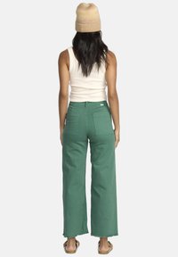 Pantaloni larghi verdi con orlo grezzo e tasche posteriori, abbinati a un top aderente beige e un cappellino di maglia chiaro.