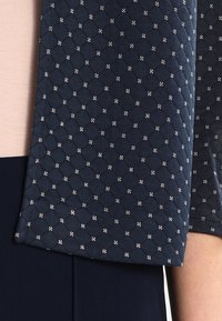 Tissu matelassé bleu marine avec motifs en losange et accents blancs. Texture douce, ourlet de manche plié. Tissu rose clair partiellement visible en dessous.