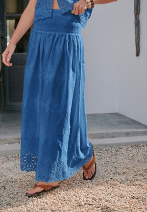 Femme portant une longue jupe bleue avec des détails en dentelle et des sandales marron, debout sur une surface de gravier près de l'entrée d'un bâtiment.