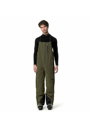 Giovane uomo che indossa pantaloni impermeabili a salopette verde oliva sopra una maglia nera a maniche lunghe, in piedi con le braccia rilassate su sfondo bianco.