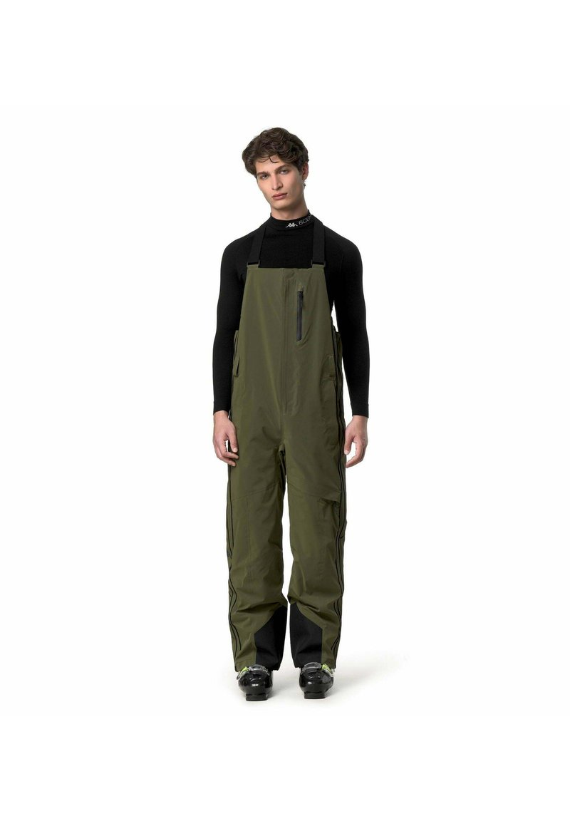 Giovane uomo che indossa pantaloni impermeabili a salopette verde oliva sopra una maglia nera a maniche lunghe, in piedi con le braccia rilassate su sfondo bianco.