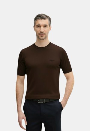 Uomo con capelli grigi che indossa una camicia aderente a maniche corte marrone scuro e pantaloni neri, in piedi con una mano in tasca su sfondo bianco.