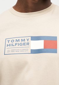 Μπεζ βαμβακερό T-shirt με μπλε και κόκκινο ορθογώνιο που περιλαμβάνει το κείμενο "TOMMY HILFIGER" και από κάτω το "ALL AMERICAN CLASSICS".