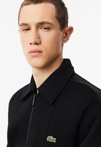 Chaqueta negra de cremallera con cuello, tela texturizada y un pequeño logo de cocodrilo verde en el lado izquierdo del pecho.