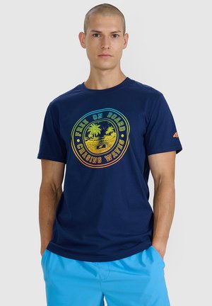 Mężczyzna o krótkich włosach nosi granatowy T-shirt z kolorowym, okrągłym tropikalnym nadrukiem oraz jasnoniebieskie szorty, ręce w kieszeniach, neutralne tło.