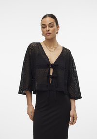 Melna mežģīņu īsā kardigāna ar V veida kakla izgriezumu, trīs ceturtdaļu piedurknēm un priekšējo saitīšanu. Iezīmējas ar sarežģītiem atvērtajiem rakstiem.