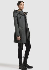 Waterdichte grijze jas met capuchon, knopen aan de voorkant, elastische taille, twee zijzakken en een gemusterde binnenvoering, gestyled met zwarte leggings en laarzen.