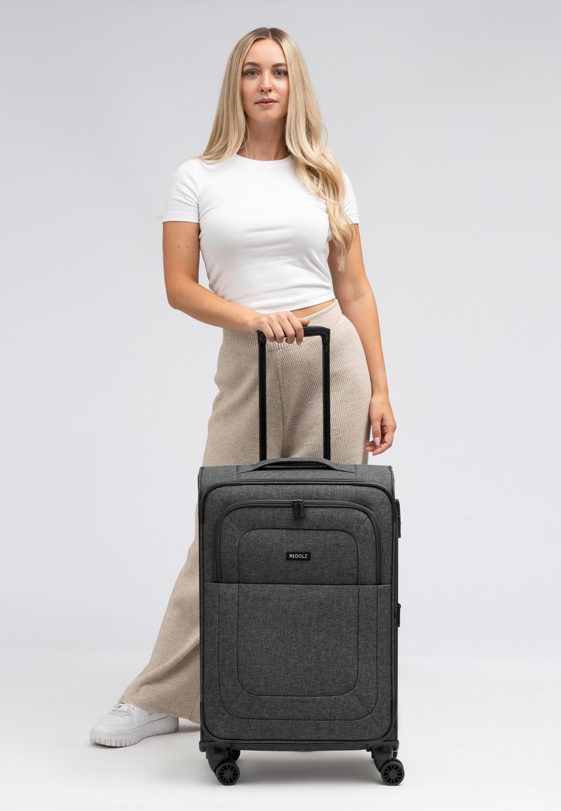 Valigia di tessuto grigio con scomparti zip, accessori neri e quattro ruote. La maniglia si estende verso l'alto per un facile trasporto.