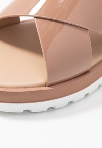 Tan patentlädersandaler med korsande remmar, en vadderad beige fotbädd och en vit gummisula med strukturerat grepp.