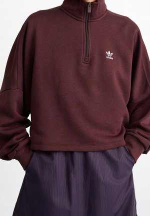 Sweat-shirt bordeaux Adidas à fermeture quart-zip avec poignets côtelés et logo sur la poitrine, porté avec un pantalon rayé sombre, mains dans les poches.