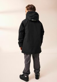 Giacca nera a maniche lunghe con cappuccio, texture liscia e marchio discreto. Abbinata a pantaloni cargo grigi e sneaker nere.