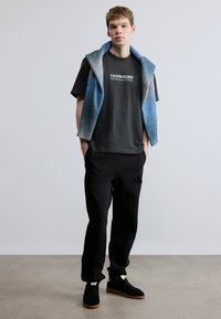 Μαύρο μπλουζάκι με τύπωμα "CALVIN KLEIN NEW YORK", συνδυασμένο με μαύρη φόρμα με λογότυπο και ένα μπλε γιλέκο με διαβάθμιση χρωμάτων που είναι ριγμένο στους ώμους.