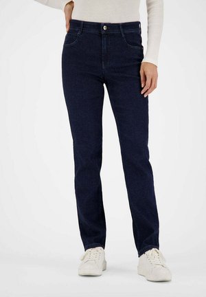 Frau trägt dunkelblaue, eng anliegende Jeans, weißes Langarmshirt und weiße Sneaker, steht mit einer Hand in der Tasche.