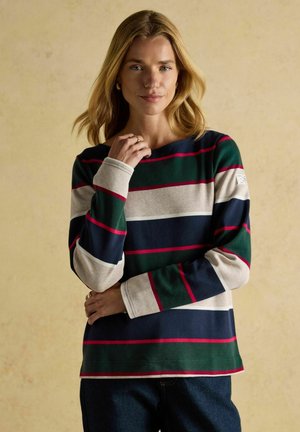 Blonde Frau in einem langärmligen Shirt mit breiten grünen, marineblauen, beigen und roten Streifen, die vor einem schlichten beigen Hintergrund steht.