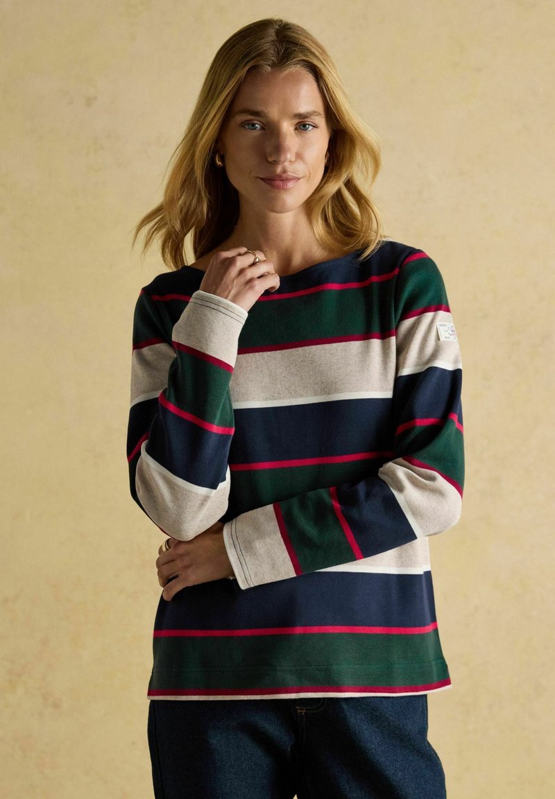 Blonde Frau in einem langärmligen Shirt mit breiten grünen, marineblauen, beigen und roten Streifen, die vor einem schlichten beigen Hintergrund steht.