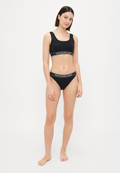 Black bralette and bikini set with a smooth texture. Features a contrasting white logo band on both pieces. Simple design, no patterns.