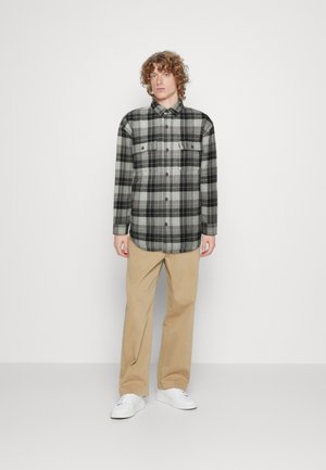 Jeune homme aux cheveux bouclés portant une chemise à carreaux noire, blanche et grise, un pantalon beige et des baskets blanches, se tenant devant un fond blanc uni.