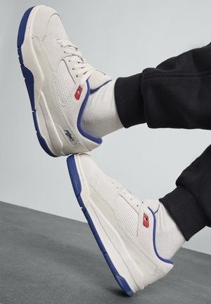 Beige semska joggesko med mesh-detaljer, blå aksenter på sålen og tungen, og rød merkevare, kombinert med grå sokker og svarte bukser.