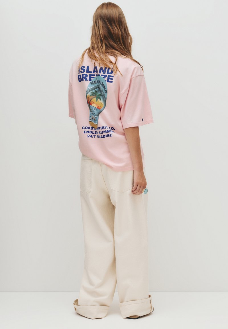 Lyserød bomulds t-shirt med et grafisk print af en strandscene på bagsiden, parret med kridhvide bukser med brede ben og omslåede bundkanter.