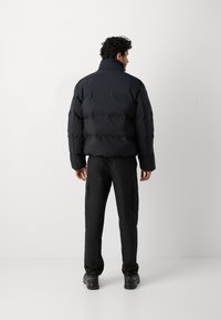 Veste matelassée noire avec des sections quiltées horizontales, col montant et manches longues, associée à un pantalon cargo noir et des chaussures sombres.