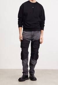 Sudadera negra de cuello redondo, combinada con pantalones cargo en negro y gris que cuentan con múltiples bolsillos y un acabado texturizado. Botas negras completan el look.